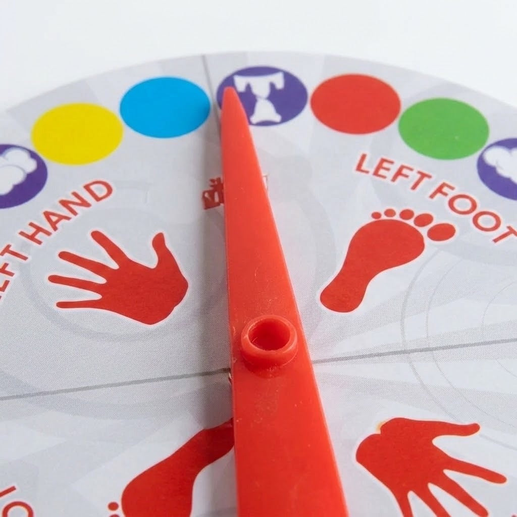 A close-up of a plastic party game spinner wheel with a red pointer. The wheel features colored circles at the top, red handprints and footprints, and text reading 'LEFT HAND' and 'LEFT FOOT'.