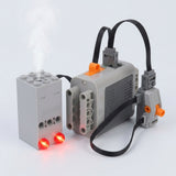 A gray plastic model exhaust sprayer connects to a battery case and orange-accented switch with black cables against a pale background.