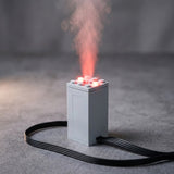 A gray plastic model exhaust sprayer stands on a textured gray surface, emitting red-lit mist with a black cable looping around its base.