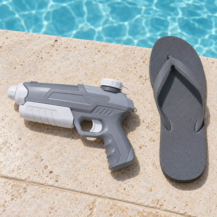 A gray and white water pistol sits beside a flip-flop on poolside stone to show its compact scale.