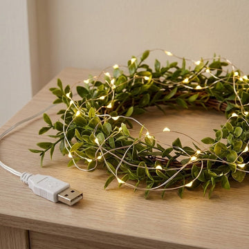 Warm white string light draped over a leafy decorative wreath on a light wooden table, with its attached USB connector visible.