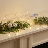 Warm white LED fairy string lights draped over green pine branches and holiday ornaments on a white mantelpiece, with a USB power adapter visible in the background.