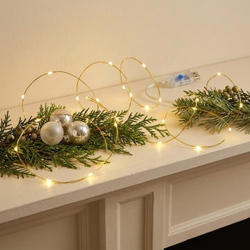 Warm white LED fairy string lights draped over green pine branches and holiday ornaments on a white mantelpiece, with a USB power adapter visible in the background.