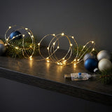 Warm white LED fairy string lights draped across a dark wooden shelf with pine branches and decorative blue, silver, and white Christmas ornaments, plus a small USB controller.