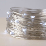 A coiled roll of silver fairy string lights with small glowing white LED bulbs, photographed on a light neutral surface against a soft gray background.