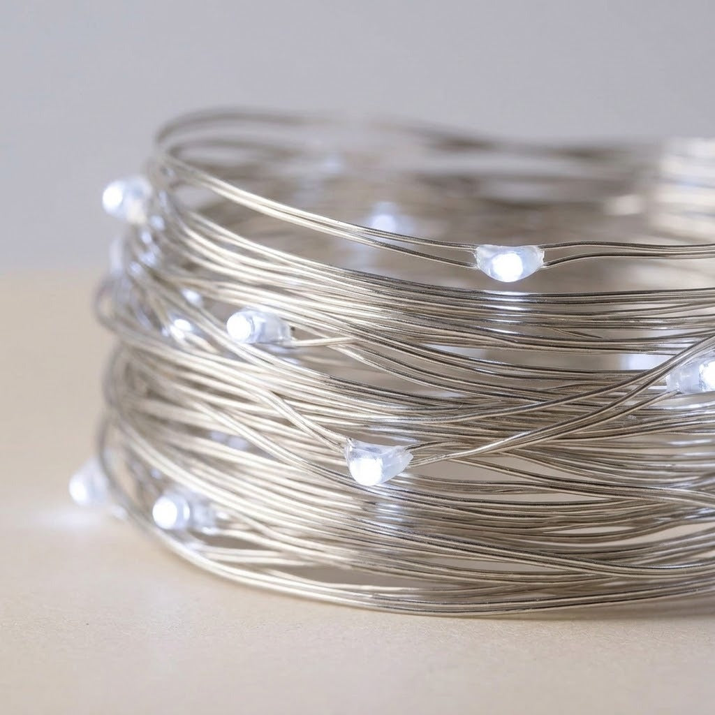 A coiled roll of silver fairy string lights with small glowing white LED bulbs, photographed on a light neutral surface against a soft gray background.