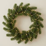 A green pine wreath decorated with warm white string lights, shown against a plain light beige wall.