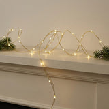 Warm white LED fairy string lights draped in loops across a white mantelpiece, flanked by green pine branches on both sides.