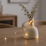 A matte cream ceramic vase with a rounded body and narrow neck, wrapped with warm fairy lights around the neck and placed on a wooden table. Dried greenery is tucked into the vase, with the string lights casting a soft glow in a cozy indoor setting.