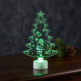 A Christmas tree shaped acrylic night light glowing green on a white base, standing on a dark wooden table.