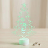 A 3D acrylic LED night light shaped like a Christmas tree with a glowing Santa Claus figure, standing on a white circular base on a light wooden surface with three small batteries nearby.