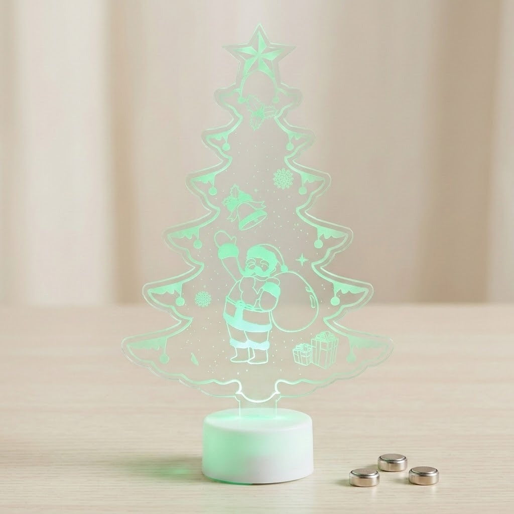 A 3D acrylic LED night light shaped like a Christmas tree with a glowing Santa Claus figure, standing on a white circular base on a light wooden surface with three small batteries nearby.