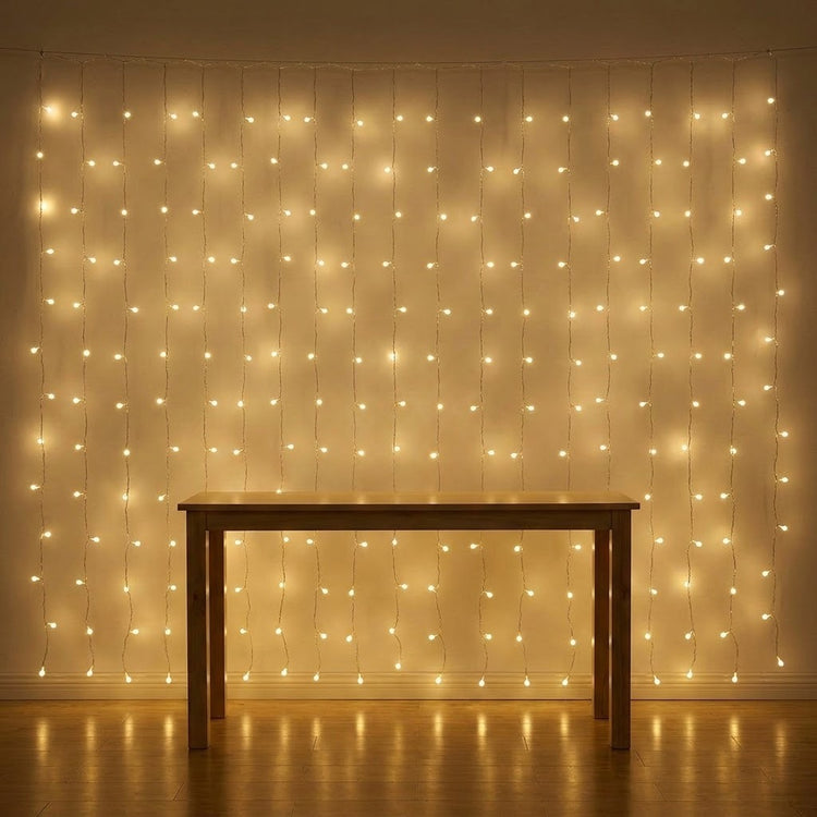 Warm white curtain string lights hanging in a grid pattern on a wall, casting a golden glow over a wooden table and floor.
