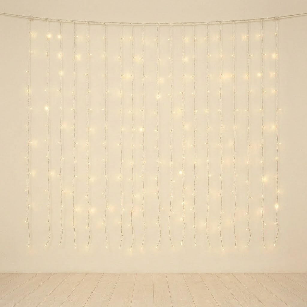 Warm white curtain string lights hanging in vertical strands against a plain off-white wall, with a light wood floor visible below.