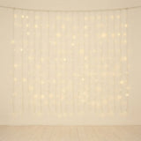 Warm white curtain string lights hanging in vertical strands against a plain off-white wall, with a light wood floor visible below.