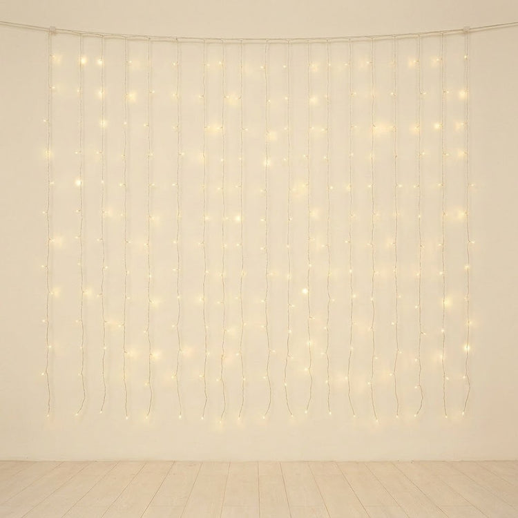 Warm white curtain string lights hanging in vertical strands against a plain off-white wall, with a light wood floor visible below.