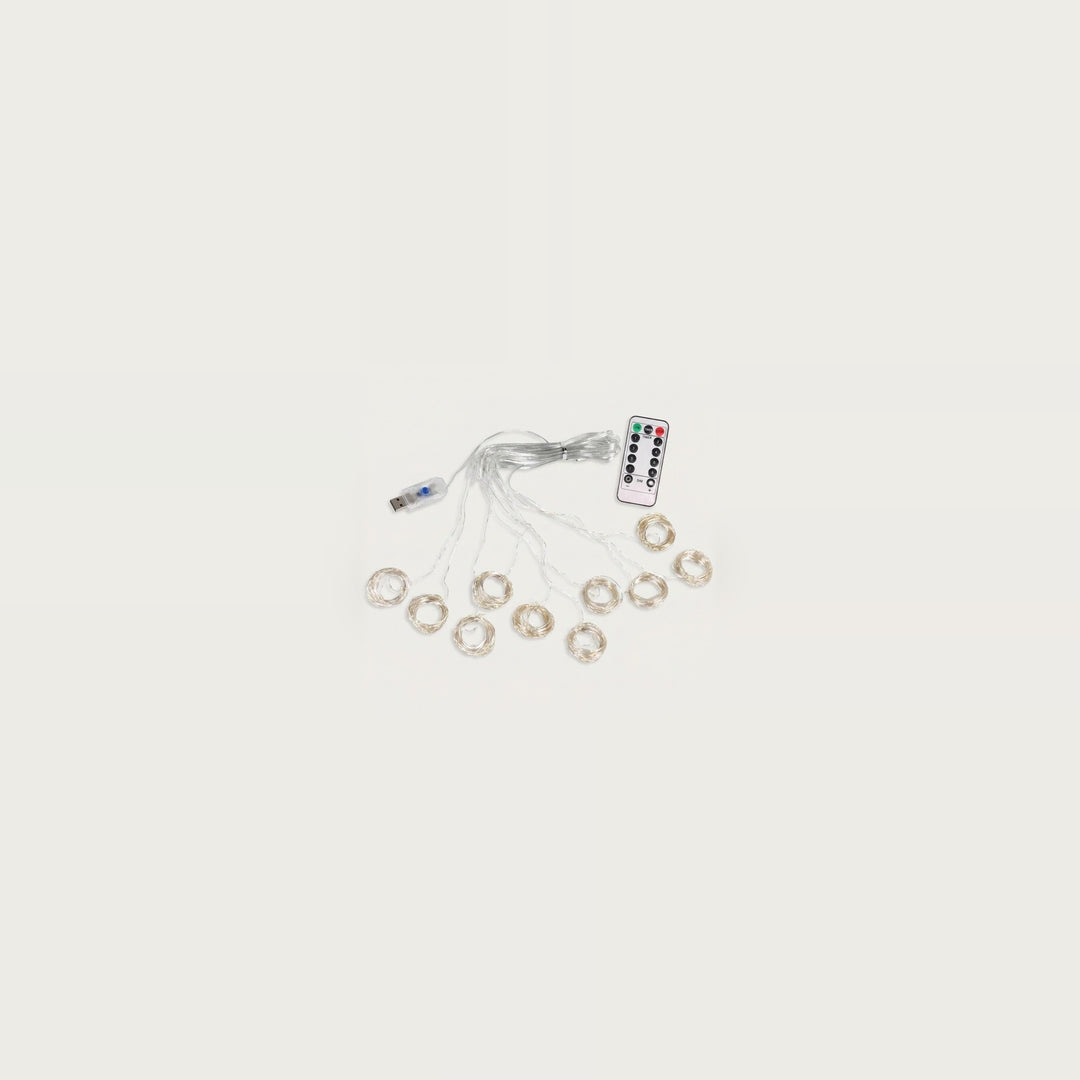 An unlit curtain light with clear wiring and small coiled strands sits beside a USB connector and white remote on an off-white background.