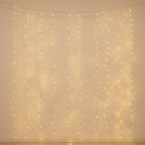 Warm white curtain string lights hanging in vertical strands against a plain beige wall, casting a soft glow.