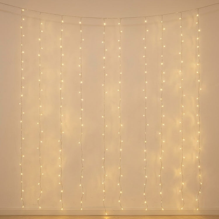 Warm white curtain string lights hanging in vertical strands against a plain beige wall, casting a soft glow.