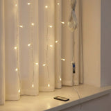Warm white curtain string lights draped over cream-colored curtains on a windowsill, with a remote control and the light cord bundle resting nearby.