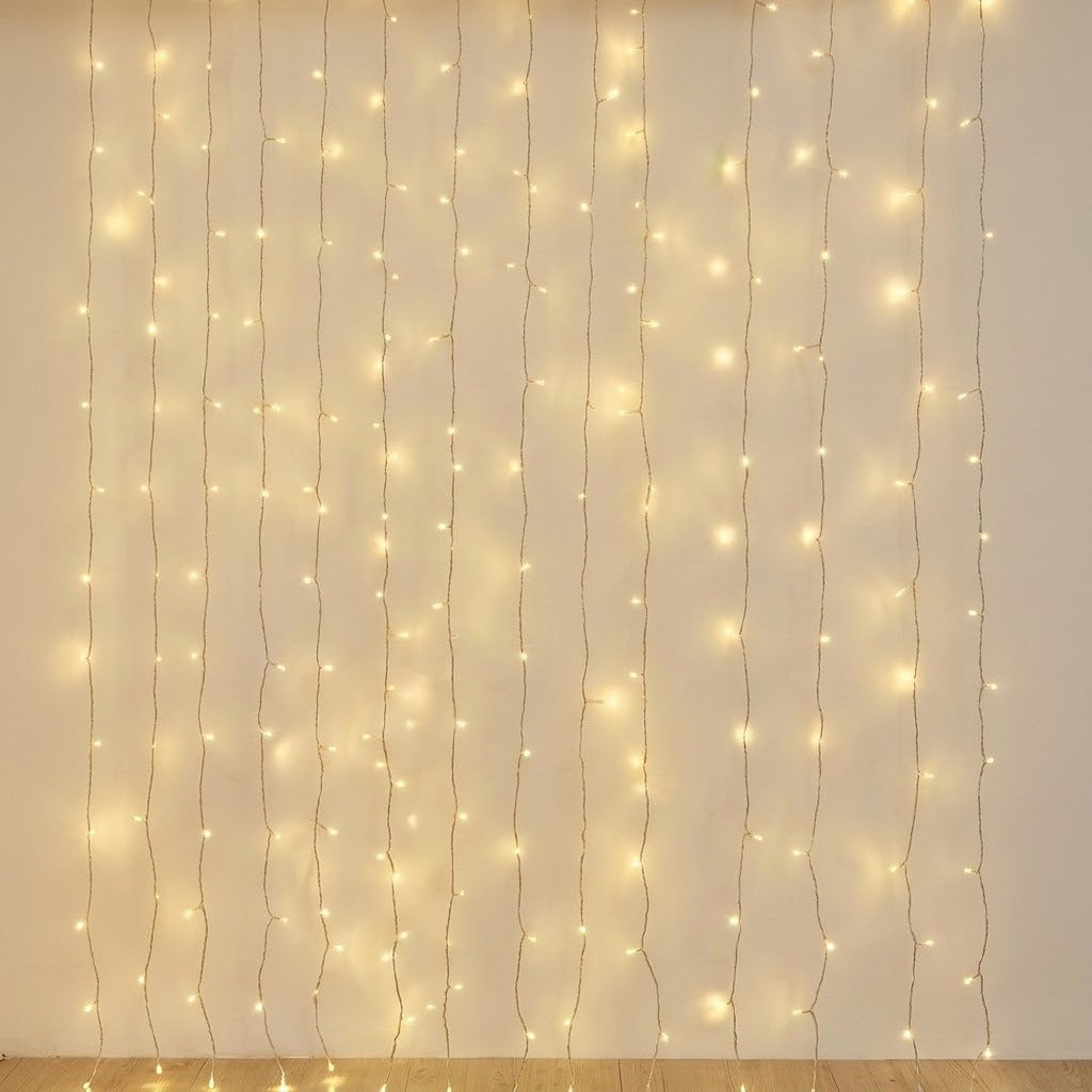 A warm white LED curtain light hangs in vertical strands against a cream wall above a wooden floor.