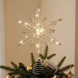 A lit silver wire star tree topper with warm white LED lights, mounted on a small striped pine tree ornament and placed among green pine branches and gold holiday decorations.