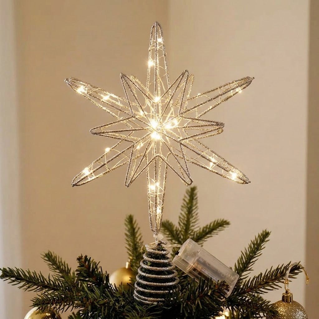 A lit silver wire star tree topper with warm white LED lights, mounted on a small striped pine tree ornament and placed among green pine branches and gold holiday decorations.