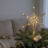A gold wire star tree topper with warm white LED lights, mounted on a green Christmas tree in a cozy living room setting.