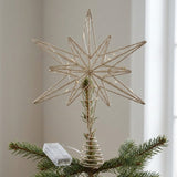 A gold glitter wire star tree topper with built-in string lights, mounted on a pine tree branch with a small clear plastic hanger visible.