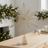 A gold wire star tree topper with a spiral gold base, shown lit with small warm white fairy lights on a light wood table in a cozy holiday room setting.