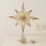 A gold wire star tree topper with warm white LED lights, mounted on a spiral spring base and shown on a tabletop with its power cord and battery pack against a plain wall.