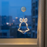 A clear acrylic Christmas bell ornament with an integrated warm white LED light and a bow detail, suspended from a suction cup on a window with a curtain next to it.