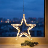 A glowing star-shaped hanging Christmas light with warm white LED bulbs, suspended in front of a window with a blurred city skyline at dusk and a pine branch below.