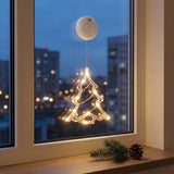A Christmas tree-shaped window string light with warm white glowing bulbs, suspended from a white circular clip attached to a window. The scene is set in a real environment with a city skyline at dusk visible outside, a wooden windowsill, and greenery and a pinecone on the sill.