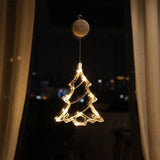 A Christmas tree-shaped LED window light with warm glowing bulbs, suspended from a circular clip against a dark window background with curtains and city lights.