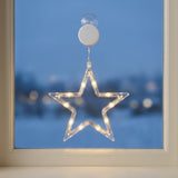 A clear star-shaped LED window light with warm white lights, suspended from a white circular suction mount, attached to a window with a snowy outdoor background visible through the glass.