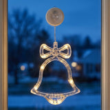 A clear plastic Christmas bell-shaped ornament with a bow, illuminated with warm white LED lights, hanging from a circular suction cup on a window with a blurred blue evening background.