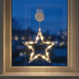 A glowing star-shaped LED window light with warm white lights, suspended from a clear suction cup on a window pane at dusk.