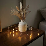 Warm white copper wire string lights loop across a wooden tabletop around a white vase of dried stems, beside a clear battery box.