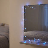 Blue LED fairy string lights draped around a wall mirror in a bedroom setting, with the mirror reflecting a bed in the background.