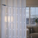 White LED curtain string lights hanging in vertical strands in front of sheer white curtains, with a power adapter visible on the left and right.