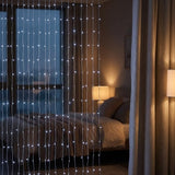 Vertical string curtain lights with small glowing bulbs hanging in front of a bedroom window, creating a soft light barrier in a dimly lit room.