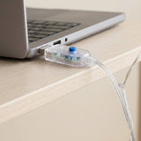 Clear USB plug with a blue button powering an LED curtain light, plugged into a laptop on a light wood desk.