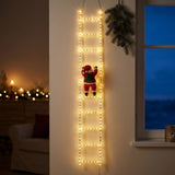 Warm white Christmas string lights form a hanging ladder with a red Santa ornament beside a window in a dim room.