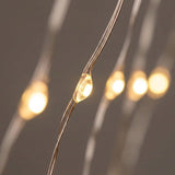 Warm white LED fairy string lights on thin copper wire, with one light bulb in sharp focus in the foreground and several blurred bulbs receding in the background against a dark gray backdrop.
