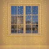 Warm white curtain string lights draped around and in front of a white-framed window, glowing against a dark blue evening sky and beige wall.
