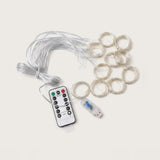 Wireless LED fairy string lights with a coiled white wire, multiple small circular light loops, a USB connector, and a white remote control with black buttons, shown on a light beige background.