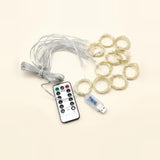 A set of fairy string lights with a coiled gray wire, multiple loops of warm white wire, a USB connector, and a white remote control with black buttons, arranged on a light beige background.