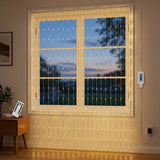 Warm white LED curtain string lights draped in vertical strands in front of a window, with a remote control on a side table and a wall-mounted controller visible in a styled room setting.