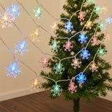 Multicolor snowflake-shaped LED string lights draped over a green Christmas tree in a room with beige walls and wood flooring.